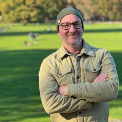 David Mitchell A headshot style image of David Mitchell standing outside in a park. He has his arms crossed and is wearing a beenie style hat.